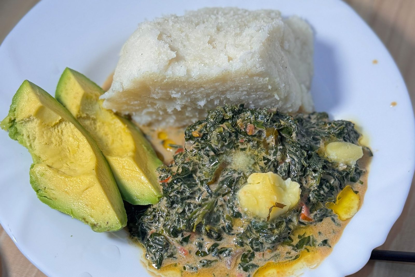 Milk greens with lumps of melting butter, served on a white plate beside a block of ugali and sliced avocado.