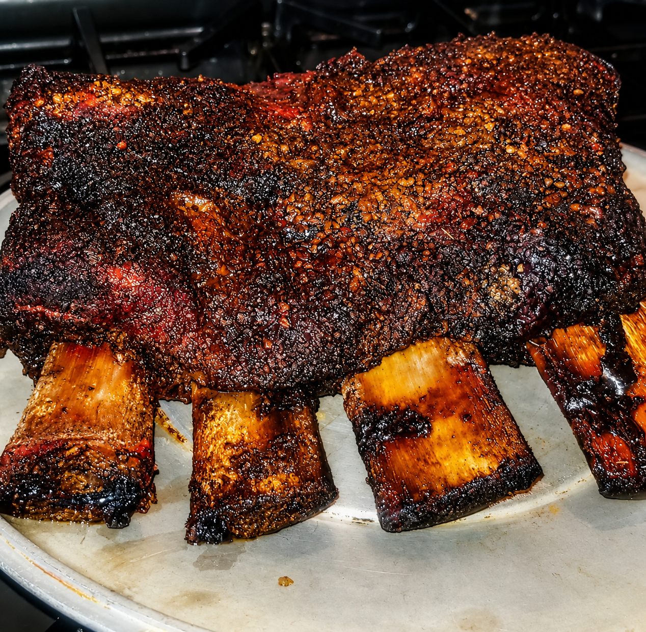 Smoked beef short ribs with a dark bark, resting on a steel pan